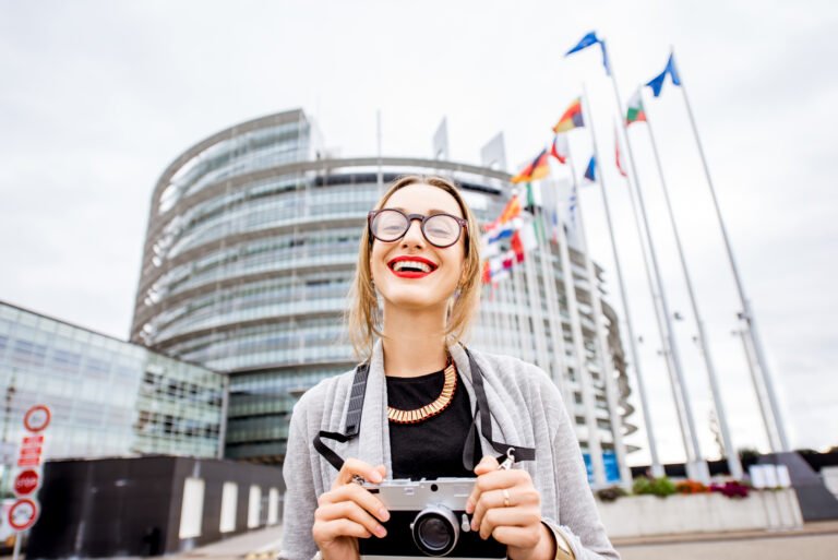 Smiling woman with a camera near the European Parliament building, symbolizing travel opportunities with a Schengen visa